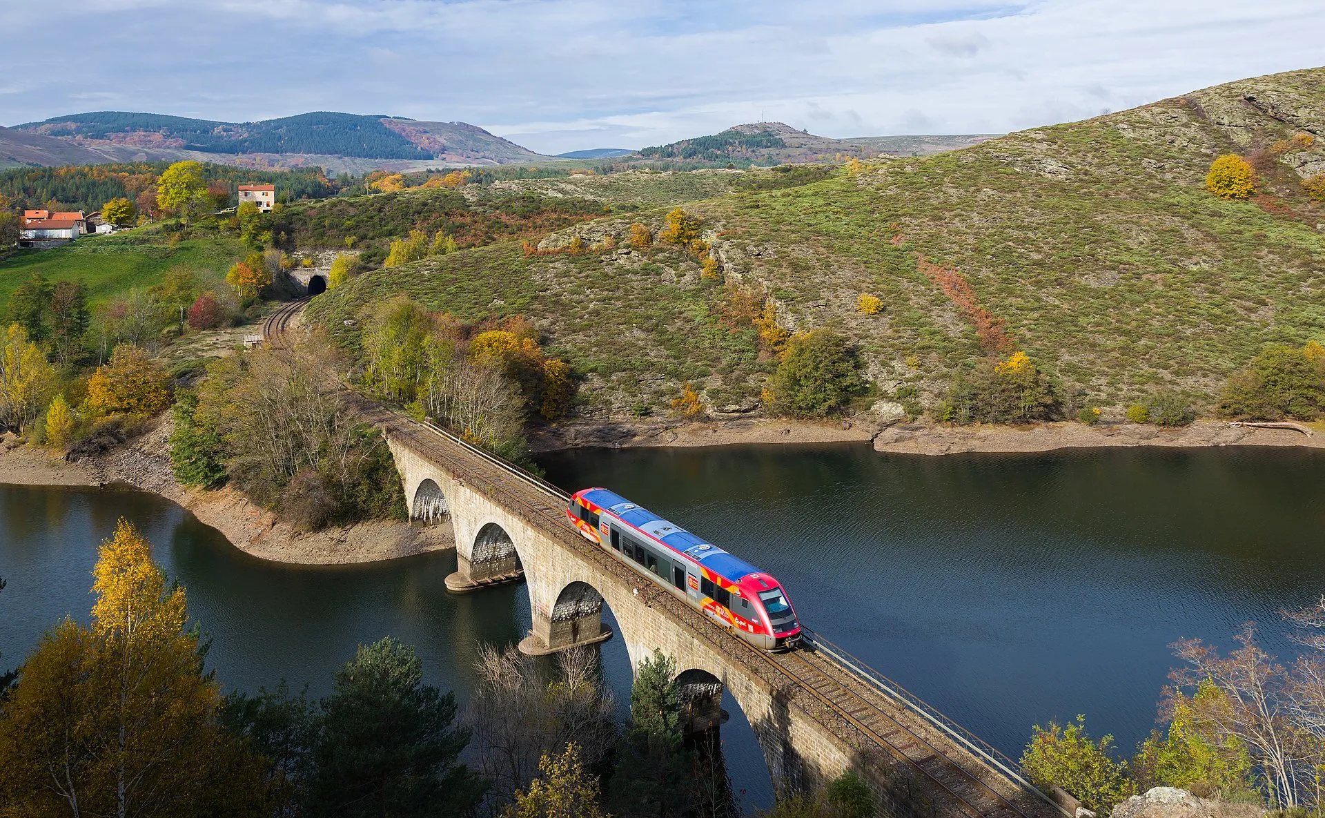 Un train régional traverse un viaduc de pierre au-dessus d’un lac, dans les collines des Cévennes en automne